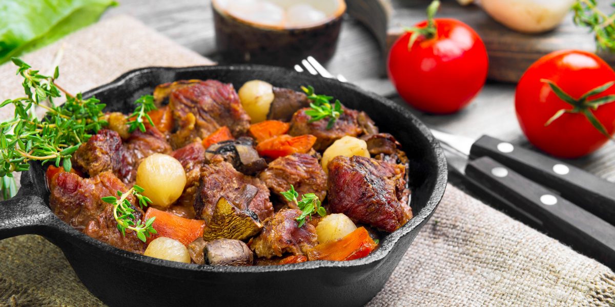 Cooking Stew meat in Burgundy (Beef Bourguignon) in cast iron frying pan with carrots, onions. Spices for Beef Bourguignon thyme, cherry tomatoes, mushrooms champignons. Gray wooden background.