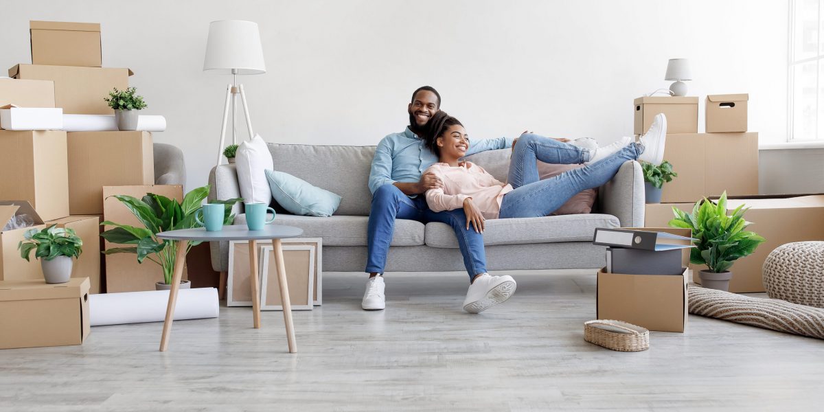 Happy glad young african american husband and wife relaxing on couch in living room with cardboard boxes and plants, enjoy moving, buying real estate, renting apartment, having rest after renovation