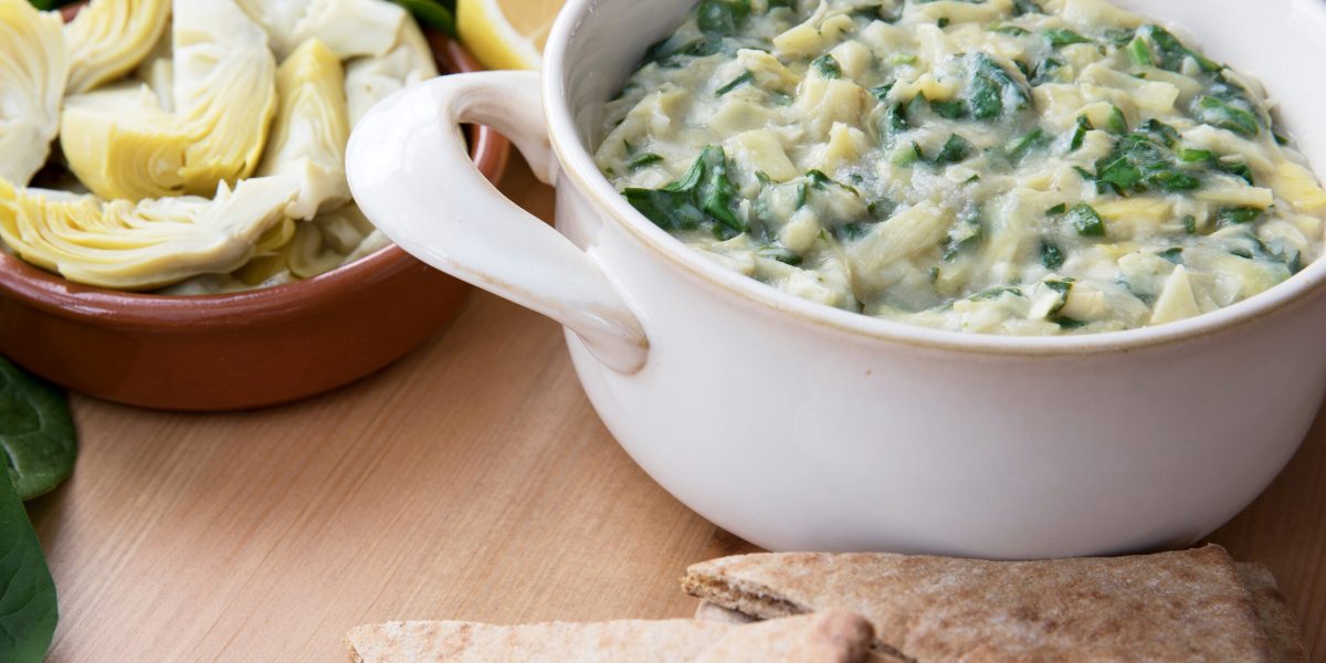 Bowl of vegan spinach artichoke dip with pita and ingredients.